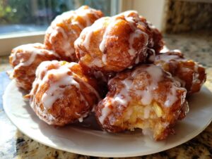 A pile of freshly fried, golden brown apple fritter pieces generously drizzled with white glaze, sitting on a white plate.