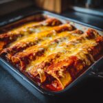 Close-up of freshly baked cheese enchiladas covered in melted orange and white cheese sauce in a dark baking dish.