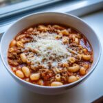 Close-up of a white bowl filled with rich pasta fagioli soup, featuring ground meat, white beans, pasta, and topped with grated Parmesan cheese.