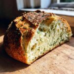Cross-section of a freshly baked artisan bread loaf showing an open crumb filled with green herbs and garlic.