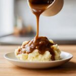 Rich homemade brown gravy being poured from a white pitcher onto a mound of fluffy mashed potatoes.