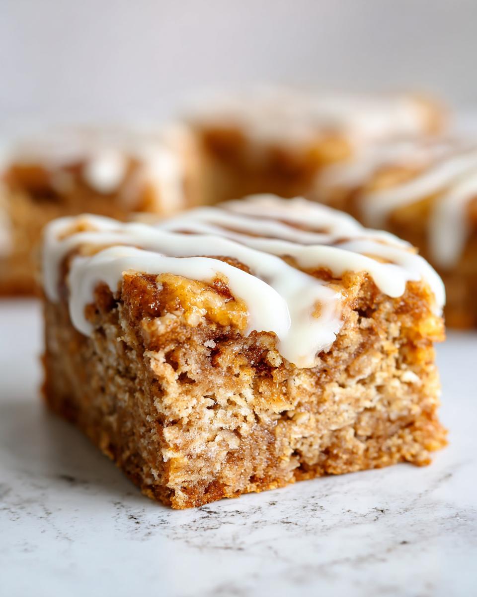 A close-up of a thick, moist square of cinnamon oatmeal bars topped with a generous drizzle of white icing.