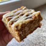A hand holding a thick, square oatmeal bars slice topped with white vanilla icing drizzle.