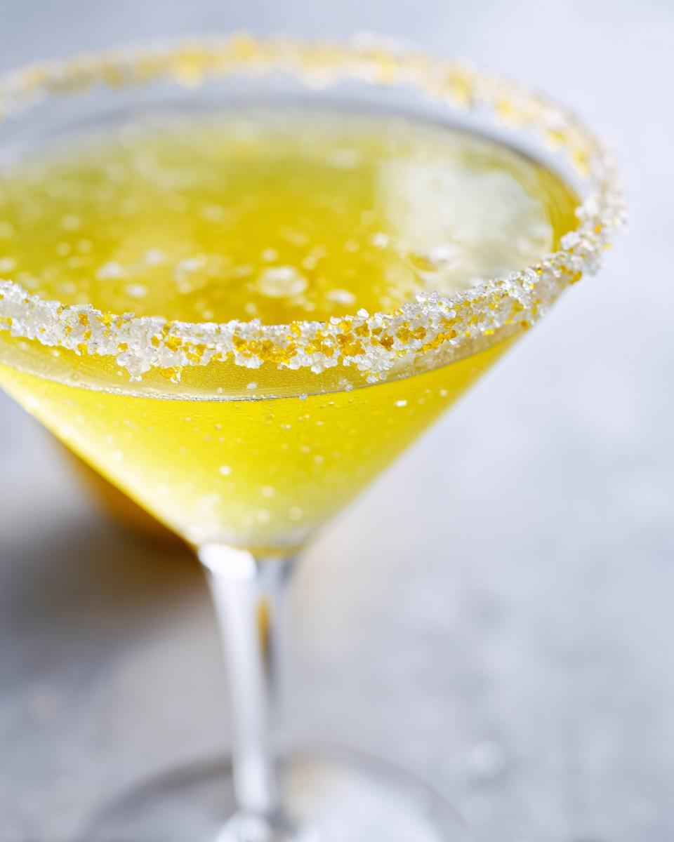 Close-up of a vibrant yellow Lemon Drop cocktail in a martini glass with a sparkling sugar and citrus zest rim.