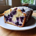 A thick slice of moist blueberry bread, bursting with blueberries and topped with a white glaze.