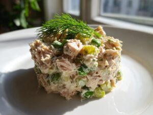 A molded portion of healthy tuna salad mixed with green peas and topped with fresh dill, served on a white plate.