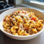 A close-up of a white bowl filled with cheesy taco pasta, featuring elbow macaroni, seasoned ground beef, and melted cheese.