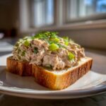 A generous scoop of healthy tuna salad, mixed with celery and topped with dill, served open-faced on toasted white bread.