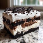 Close-up of a layered ice cream cake slice featuring dark chocolate cookie layers, white cream, and topped with whole Oreos.