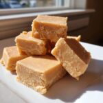 A close-up stack of creamy, light brown peanut butter fudge squares resting on a white plate.