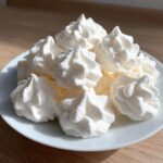 A close-up of several perfectly piped white meringue cookies piled on a small white plate.