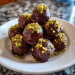 A close-up of stacked, glossy, chocolate-covered dubai chocolate balls topped with bright green crushed pistachios.