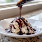 A spoonful of rich chocolate syrup being poured over scoops of vanilla ice cream, creating delicious drizzles.