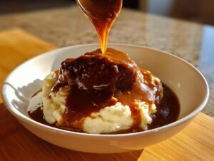 A spoonful of rich homemade brown gravy is being poured over a mound of mashed potatoes in a white bowl.