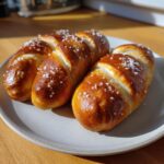Close-up of three shiny, golden brown pretzel dogs buns topped with coarse salt crystals.