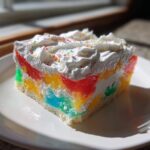 A colorful slice of jello poke cake featuring layers of rainbow gelatin, white cake, and topped with whipped cream and sprinkles.