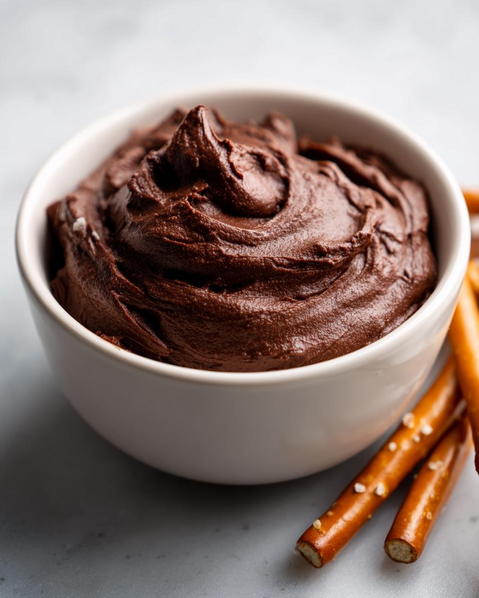 A close-up of thick, rich chocolate dip swirled in a white bowl, served alongside salted pretzel sticks.