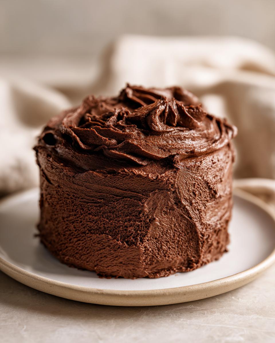 A small, round cake generously covered in thick, rich chocolate frosting, ready to serve.