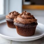 Two dark chocolate cupcakes topped with thick, rich chocolate frosting sitting on a white plate.