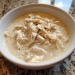 A close-up of a white bowl filled with thick, creamy cream of chicken soup topped generously with shredded chicken.