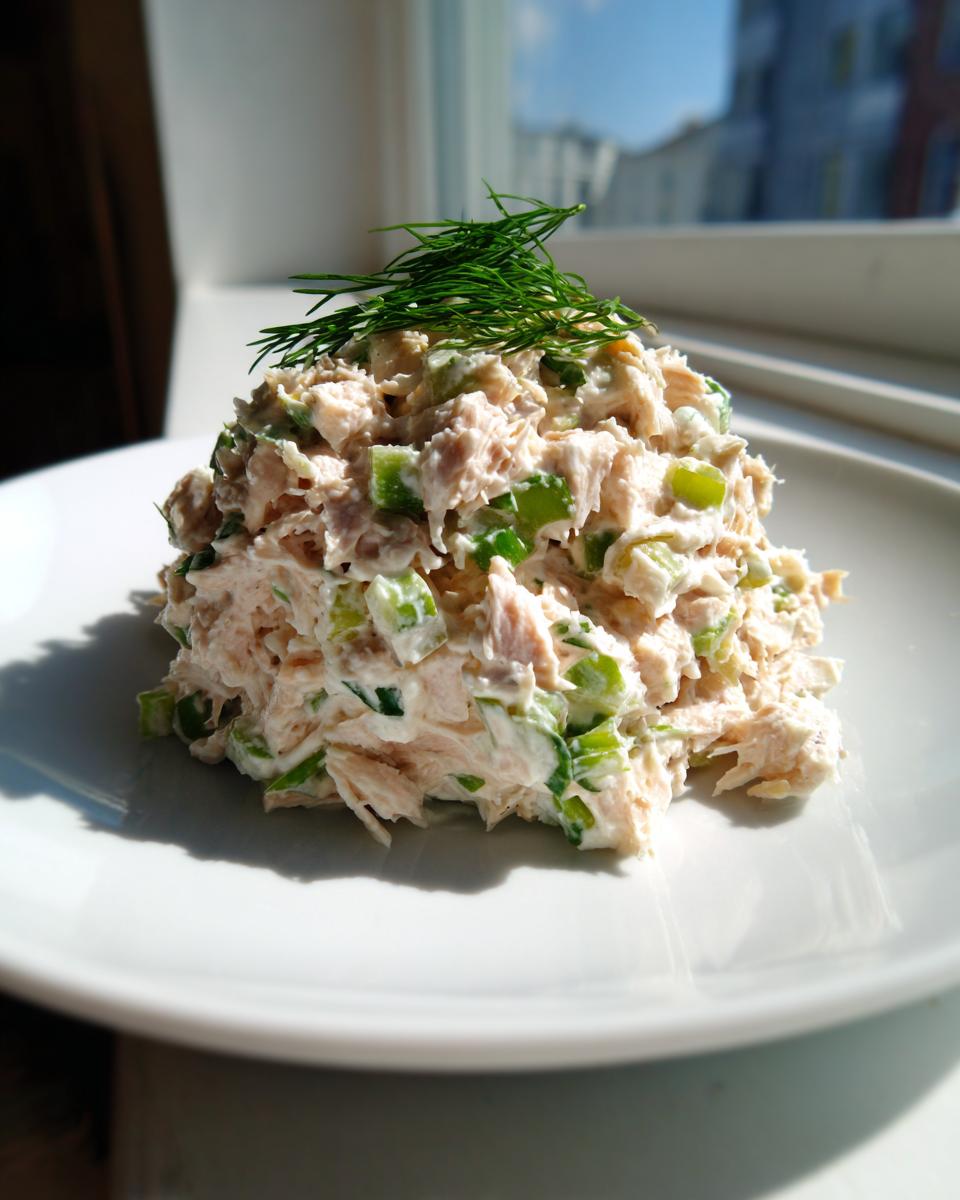 A mound of creamy, chunky healthy tuna salad mixed with green celery, topped with fresh dill, served on a white plate.