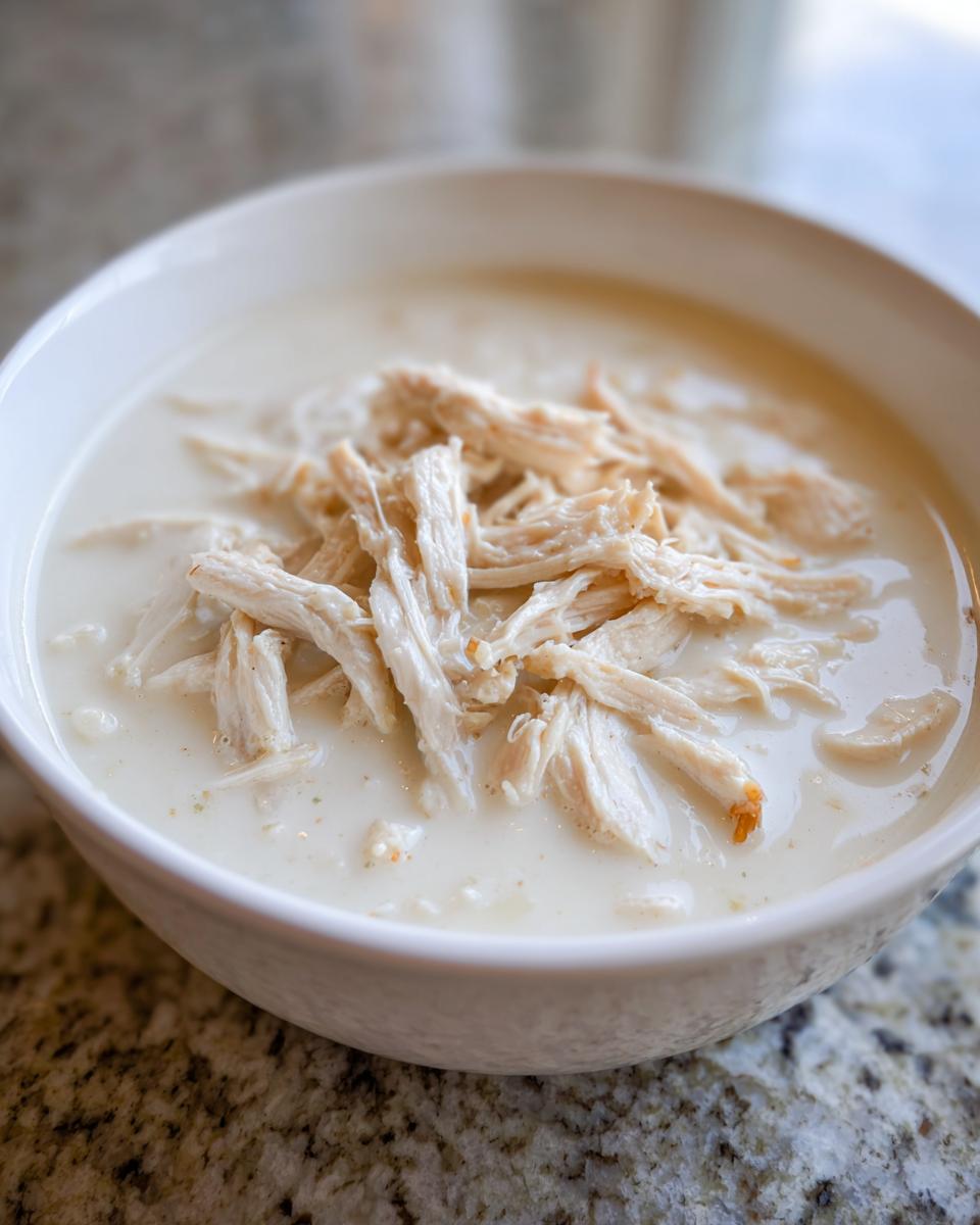 Close-up of a white bowl filled with creamy liquid and topped generously with shredded chicken, representing cream of chicken soup.