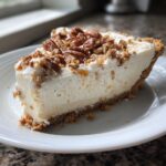 A thick slice of creamy million dollar pie topped generously with pecans and crumbs on a white plate.