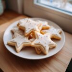 A plate piled high with star-shaped vegan sugar cookies topped with white icing and small white sprinkles.