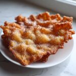 Close-up of crispy, golden-brown buñuelos generously coated in sparkling sugar, stacked on a white plate.