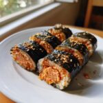 Six slices of homemade tuna kimbap arranged on a white plate, featuring a rich orange tuna filling and sprinkled with sesame seeds.