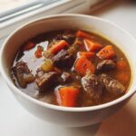 Close-up of a steaming bowl of rich vegetable beef soup with large chunks of beef and bright orange carrots.