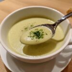 A spoonful of creamy, light green celery soup garnished with fresh parsley being lifted from a white bowl.