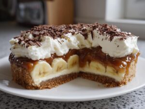 A cross-section view of an amazing banoffee pie showing layers of biscuit base, cream, bananas, toffee, and whipped cream topping with chocolate shavings.