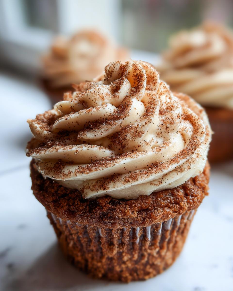 A close-up of a single churro cupcake topped with swirled cinnamon frosting and dusted heavily with cinnamon sugar.