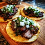 Three delicious steak tacos topped with diced white onions and cilantro resting on a wooden cutting board.