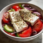 Close-up of a vibrant greek salad featuring large blocks of feta cheese seasoned with oregano, tomatoes, cucumbers, and olives.