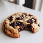A close-up of a chewy brown butter chocolate chip cookie with melted chocolate chunks and flaky sea salt, featuring a bite taken out.