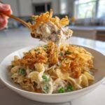 A golden spoon lifts a cheesy, creamy scoop of tuna noodle casserole topped with crispy onions.