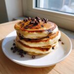 A tall stack of three fluffy chocolate chip pancakes topped with mini chocolate chips.