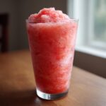 Close-up of a tall glass filled with a bright red, icy strawberry daiquiri, sweating condensation.