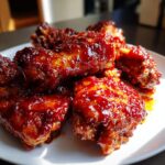 Close-up of sticky, glossy gochujang chicken wings piled on a white plate, catching the sunlight.