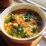 A close-up view of a steaming bowl of italian orzo spinach soup, featuring orzo pasta, dark green spinach, and diced carrots.