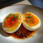 Two halves of soft-boiled marinated eggs with vibrant orange yolks, topped with scallions and sesame seeds.