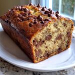 A close-up of a freshly baked, moist chocolate chip banana bread loaf with melted chips on top, resting on a white plate.