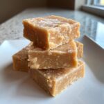A stack of four creamy squares of peanut butter fudge recipe dusted lightly with powdered sugar on a white plate.