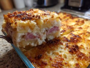 A fork lifts a creamy square portion of scalloped potatoes and ham from a baking dish with a golden-brown, bubbly crust.