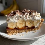 A generous slice of banoffee pie featuring a crumbly base, thick caramel, banana slices, whipped cream, and chocolate shavings.
