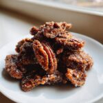 A pile of freshly made candied pecans coated heavily in sparkling sugar crystals, resting on a white plate.