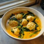 A close-up of a white bowl filled with vibrant yellow coconut curry soup, featuring several white dumplings and wilted green spinach.