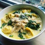 A close-up of a bowl of creamy chicken tortellini soup topped with shredded chicken, spinach, and grated Parmesan cheese.
